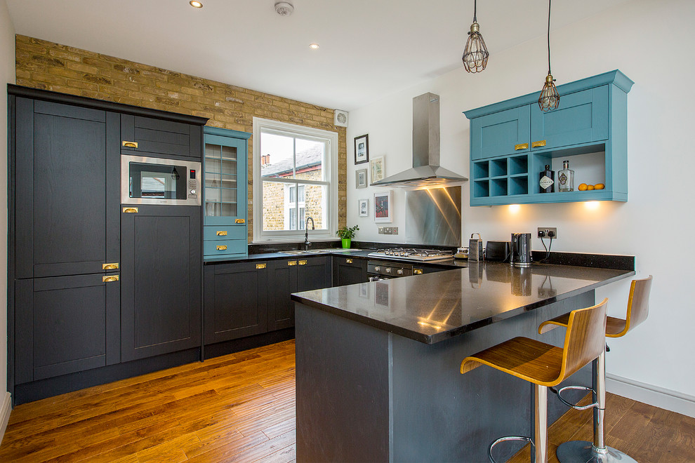 Photo of a mid-sized transitional u-shaped kitchen in London with a double-bowl sink, metallic splashback, stainless steel appliances, medium hardwood floors, a peninsula, brown floor, black benchtop, shaker cabinets and blue cabinets.