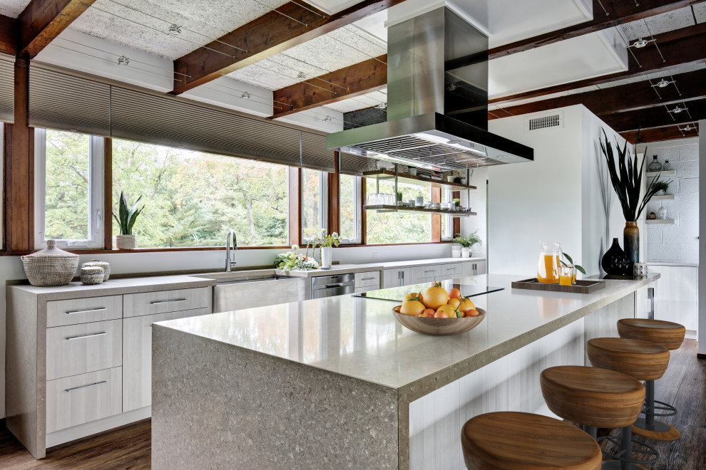 This is an example of a small urban galley open plan kitchen in New York with a belfast sink, flat-panel cabinets, grey cabinets, engineered stone countertops, stainless steel appliances, medium hardwood flooring, an island, brown floors, grey worktops and exposed beams.
