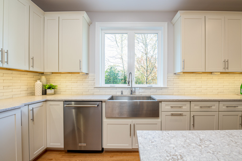 Country kitchen in DC Metro with a farmhouse sink, recessed-panel cabinets, white cabinets, quartz benchtops, grey splashback, stainless steel appliances, light hardwood floors and with island.