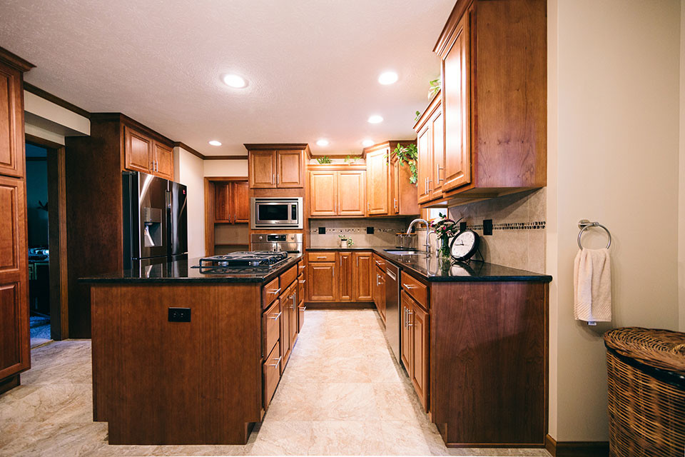 This is an example of a traditional l-shaped eat-in kitchen in Other with an undermount sink, raised-panel cabinets, medium wood cabinets, quartz benchtops, beige splashback, porcelain splashback, stainless steel appliances, porcelain floors, with island, beige floor and black benchtop.