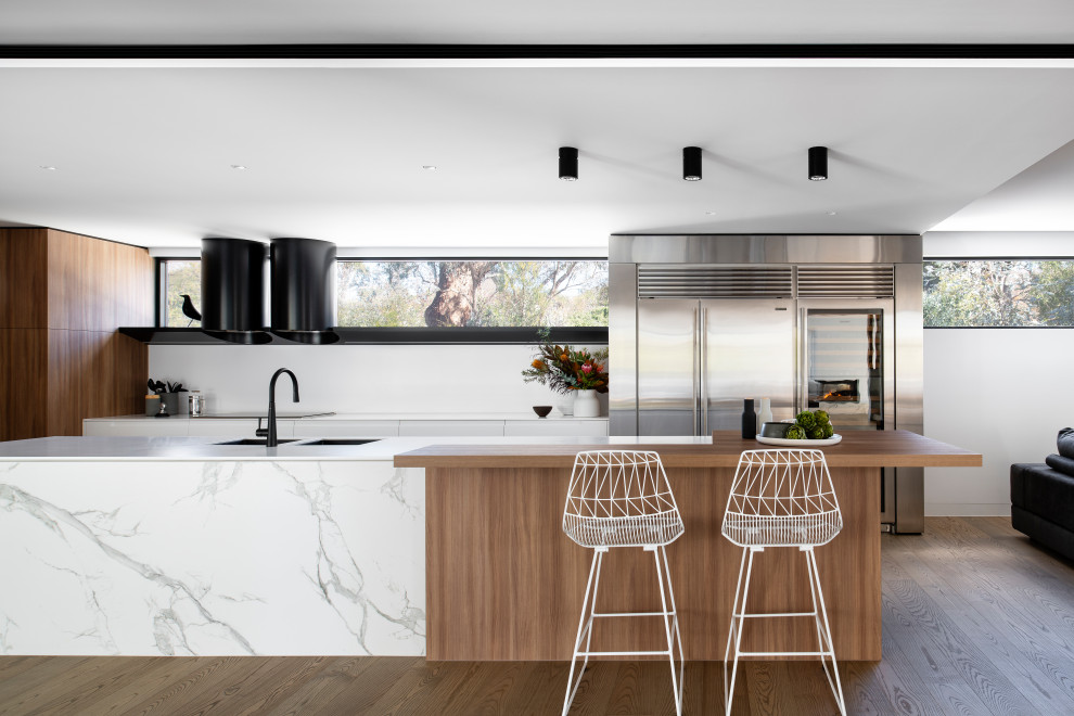 Example of a large trendy l-shaped medium tone wood floor kitchen pantry design in Sydney with an undermount sink, flat-panel cabinets, solid surface countertops, white backsplash, black appliances, an island and white countertops