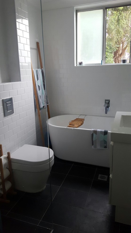 Photo of a small contemporary master bathroom in Sydney with furniture-like cabinets, white cabinets, a freestanding tub, a corner shower, a wall-mount toilet, white tile, ceramic tile, white walls, slate floors, an integrated sink, laminate benchtops, black floor and a hinged shower door.