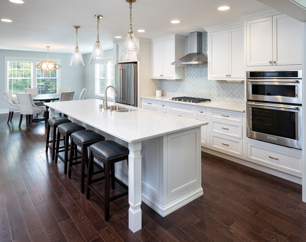 Photo of a mid-sized transitional l-shaped eat-in kitchen in Minneapolis with a double-bowl sink, recessed-panel cabinets, white cabinets, marble benchtops, grey splashback, ceramic splashback, stainless steel appliances, dark hardwood floors, with island and brown floor.