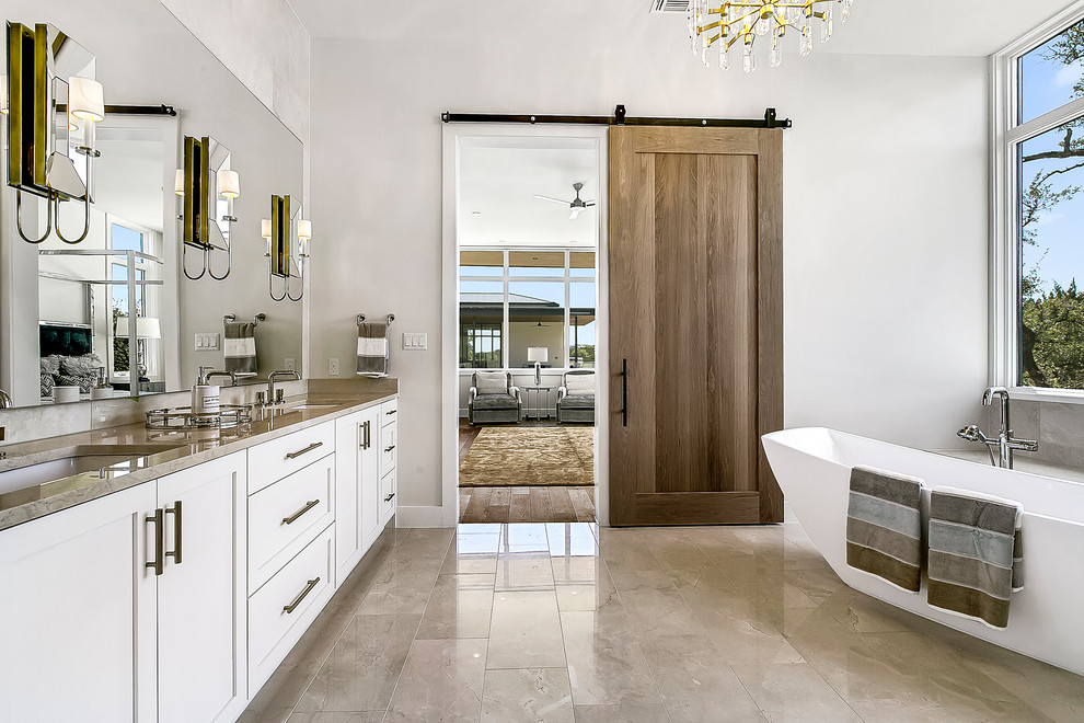 This is an example of a large contemporary master bathroom in Austin with shaker cabinets, white cabinets, a freestanding tub, an alcove shower, gray tile, marble, white walls, porcelain floors, an undermount sink, marble benchtops, beige floor, a hinged shower door and black benchtops.