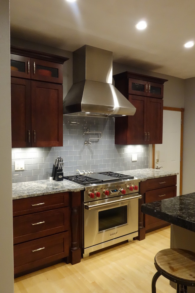 Eat-in kitchen - mid-sized light wood floor eat-in kitchen idea with a farmhouse sink, recessed-panel cabinets, dark wood cabinets, granite countertops, gray backsplash, glass tile backsplash, stainless steel appliances and an island