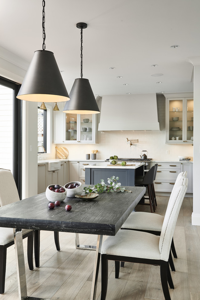 Mid-sized transitional u-shaped light wood floor and beige floor kitchen/dining room combo photo with a farmhouse sink, shaker cabinets, white cabinets, quartzite countertops, white backsplash, ceramic backsplash, stainless steel appliances, an island, white countertops and white walls