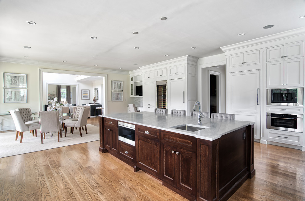 Open concept kitchen - huge transitional u-shaped light wood floor and brown floor open concept kitchen idea in New York with a farmhouse sink, shaker cabinets, white cabinets, quartzite countertops, white backsplash, porcelain backsplash, paneled appliances, an island and multicolored countertops