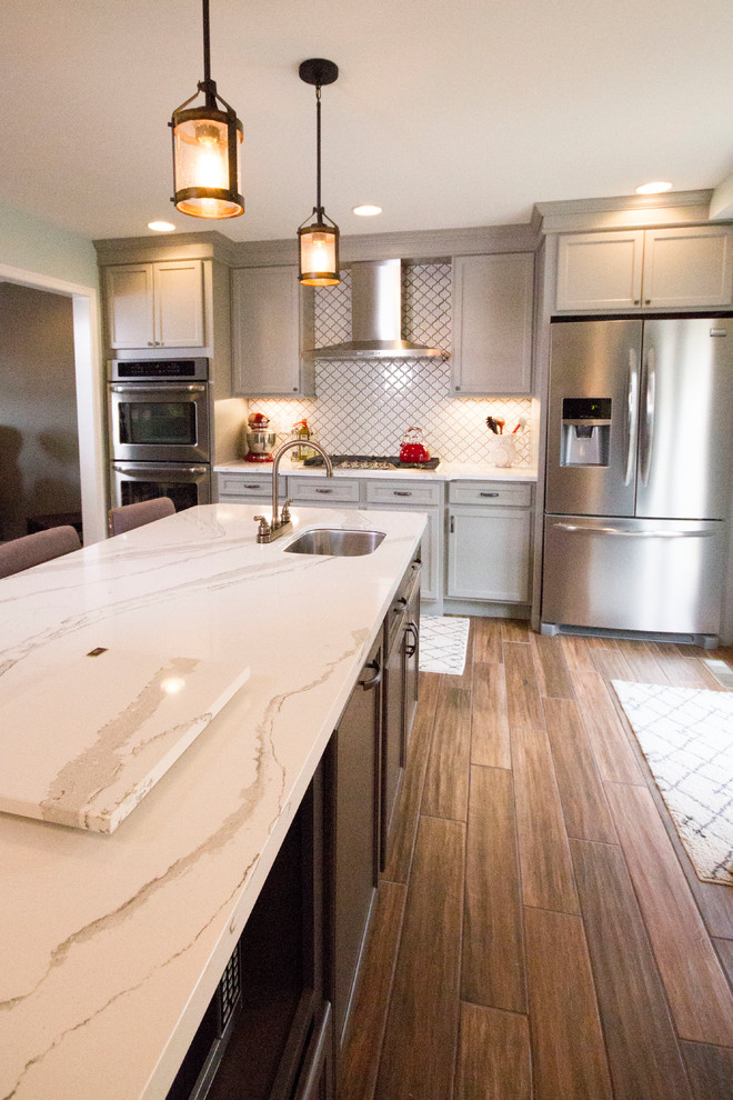 Enclosed kitchen - large transitional l-shaped medium tone wood floor and brown floor enclosed kitchen idea in Cincinnati with a farmhouse sink, recessed-panel cabinets, gray cabinets, marble countertops, white backsplash, porcelain backsplash, stainless steel appliances and an island