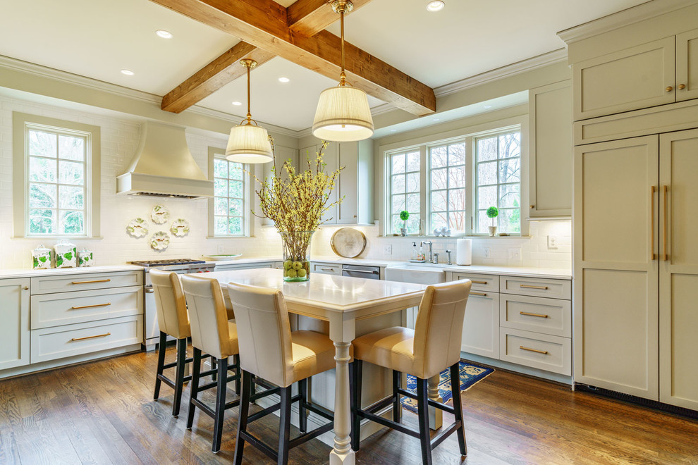 This is an example of a traditional l-shaped kitchen in Other with a farmhouse sink, shaker cabinets, beige cabinets, white splashback, panelled appliances, medium hardwood floors, with island, brown floor and white benchtop.