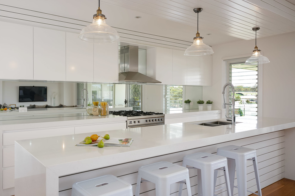 Beach style galley kitchen in Other with a double-bowl sink, flat-panel cabinets, white cabinets, mirror splashback, stainless steel appliances, a peninsula and white benchtop.