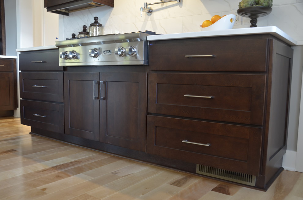 Large transitional u-shaped light wood floor home design photo with an undermount sink, shaker cabinets, dark wood cabinets, quartz countertops, white backsplash, stainless steel appliances, an island and stone tile backsplash