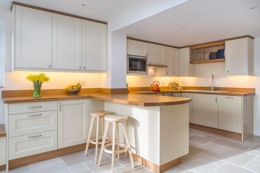 Design ideas for a transitional l-shaped kitchen in Oxfordshire with wood benchtops, travertine floors, a drop-in sink, shaker cabinets, beige cabinets and a peninsula.