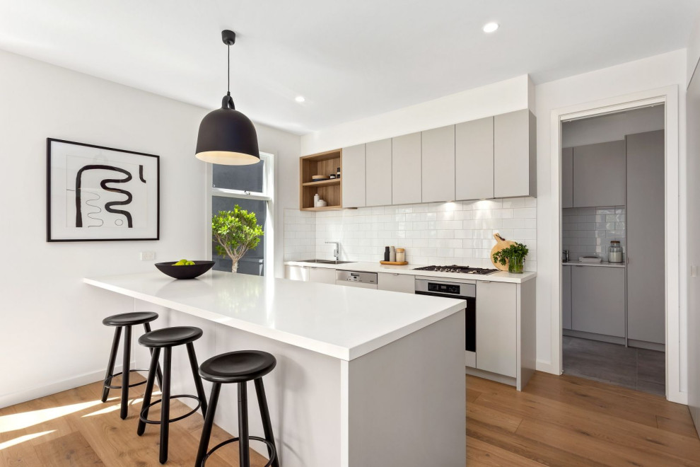 Design ideas for a contemporary galley kitchen in Melbourne with flat-panel cabinets, grey cabinets, white splashback, subway tile splashback, stainless steel appliances, medium hardwood floors, a peninsula, brown floor and white benchtop.