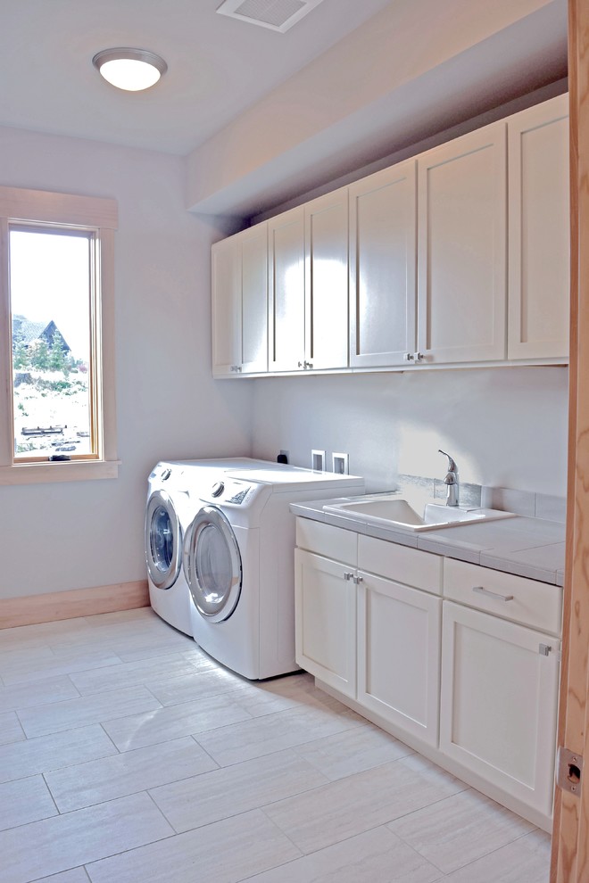 Large traditional single-wall utility room in Seattle with a drop-in sink, shaker cabinets, white cabinets, tile benchtops, grey walls, porcelain floors, a side-by-side washer and dryer and grey benchtop.