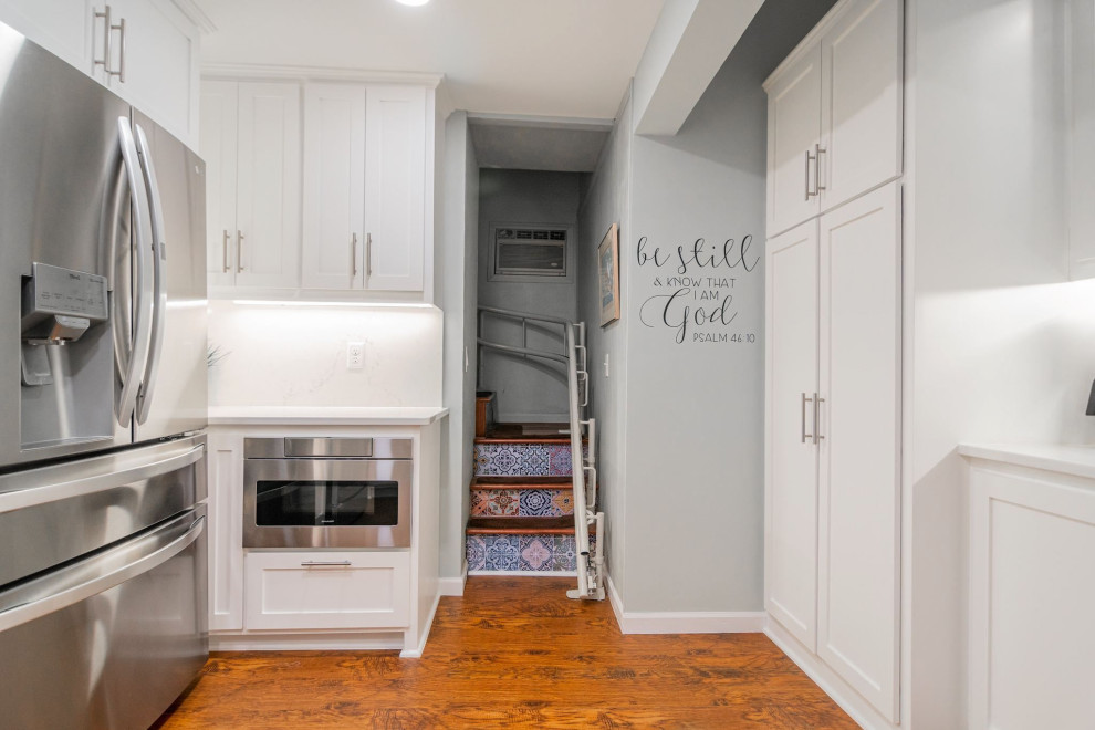 Small transitional u-shaped separate kitchen in Minneapolis with a farmhouse sink, shaker cabinets, white cabinets, quartz benchtops, white splashback, engineered quartz splashback, stainless steel appliances, vinyl floors, no island, brown floor and white benchtop.
