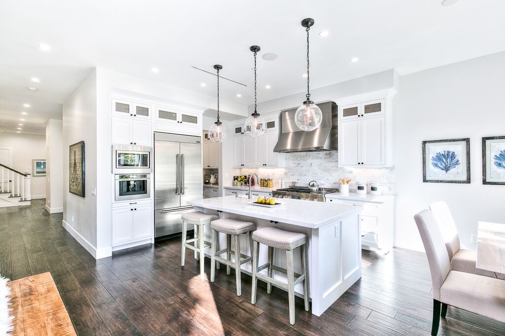 Large transitional l-shaped eat-in kitchen in Orange County with a farmhouse sink, shaker cabinets, white cabinets, marble benchtops, white splashback, marble splashback, stainless steel appliances, dark hardwood floors, with island and brown floor.