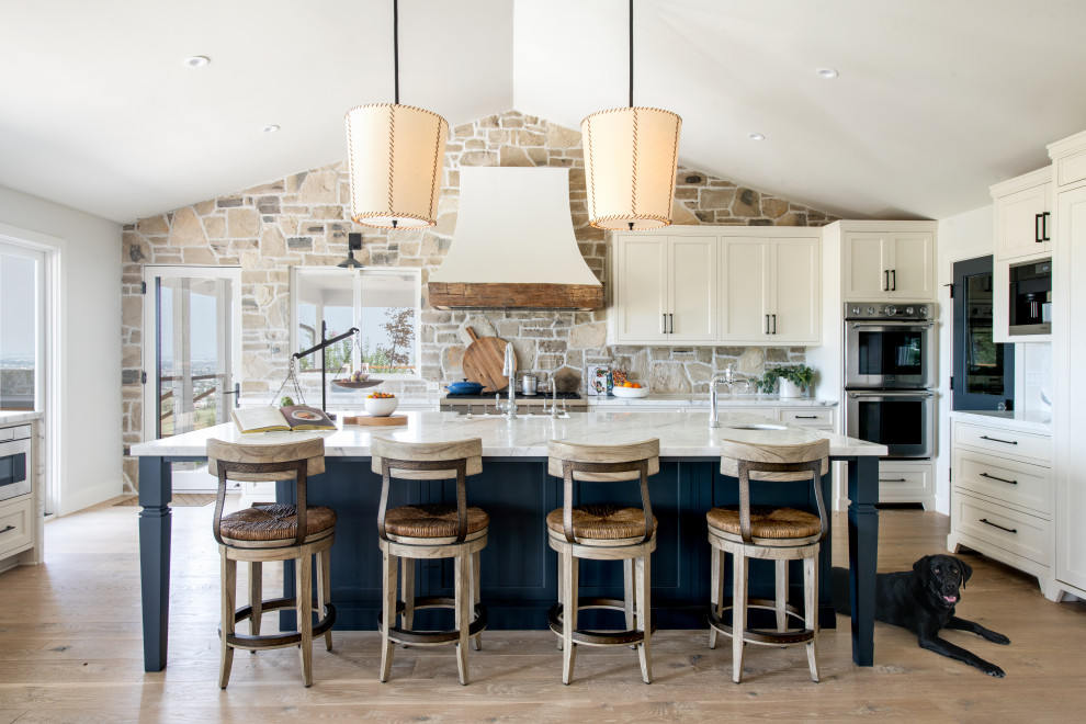 Design ideas for a country u-shaped kitchen in Los Angeles with shaker cabinets, white cabinets, stone tile splashback, stainless steel appliances, light hardwood floors, with island, white benchtop and vaulted.