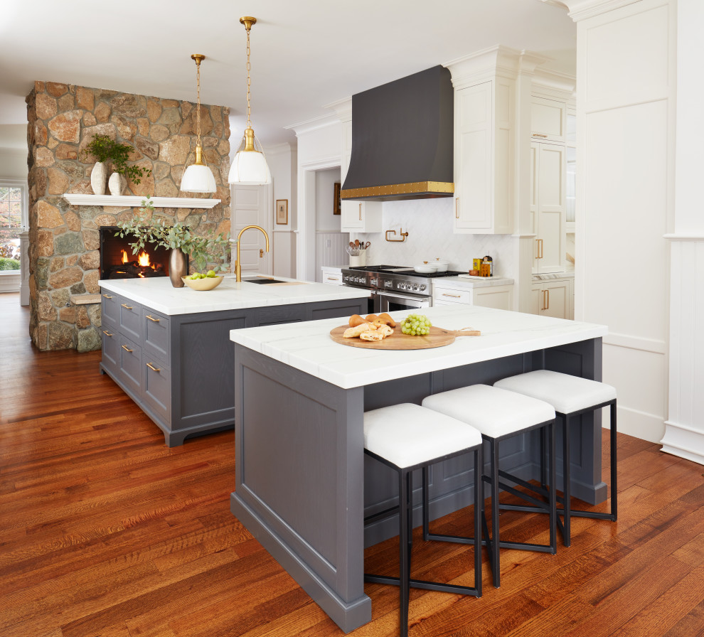Large traditional u-shaped open plan kitchen in New York with a farmhouse sink, shaker cabinets, white cabinets, marble benchtops, white splashback, marble splashback, stainless steel appliances, medium hardwood floors, multiple islands, brown floor and white benchtop.