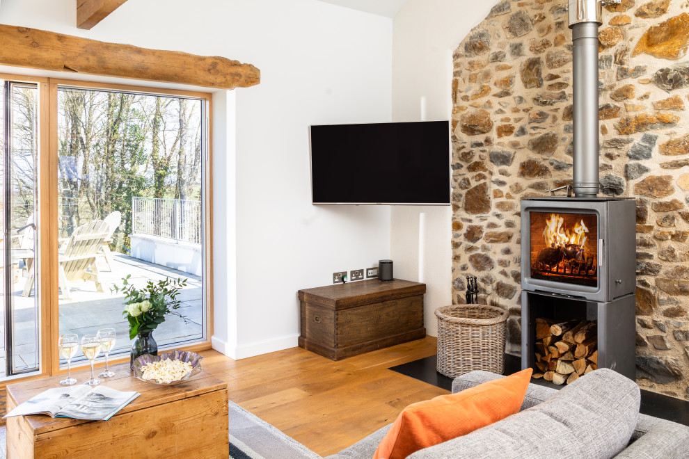 Landhausstil Wohnzimmer mit weißer Wandfarbe, braunem Holzboden, Kaminofen, Kaminumrandung aus Stein, TV-Wand und braunem Boden in Cornwall
