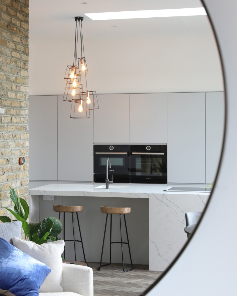 Design ideas for a medium sized contemporary grey and white single-wall open plan kitchen in London with a built-in sink, flat-panel cabinets, grey cabinets, marble worktops, black appliances, dark hardwood flooring, an island, brown floors, white worktops and feature lighting.