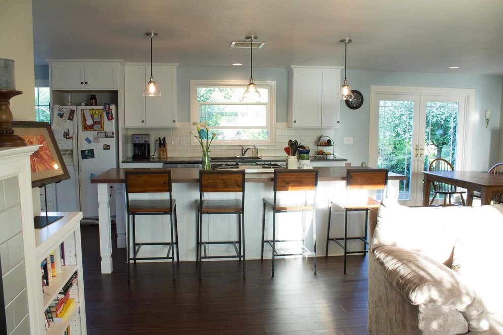 Example of a mid-sized transitional galley dark wood floor and brown floor open concept kitchen design with an undermount sink, shaker cabinets, white cabinets, wood countertops, white backsplash, porcelain backsplash, white appliances and an island