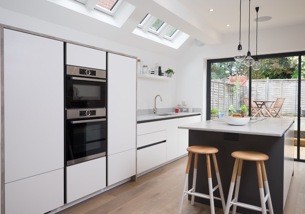 This is an example of a large contemporary single-wall open plan kitchen in Hampshire with an undermount sink, flat-panel cabinets, white cabinets, white splashback, stainless steel appliances, with island and light hardwood floors.