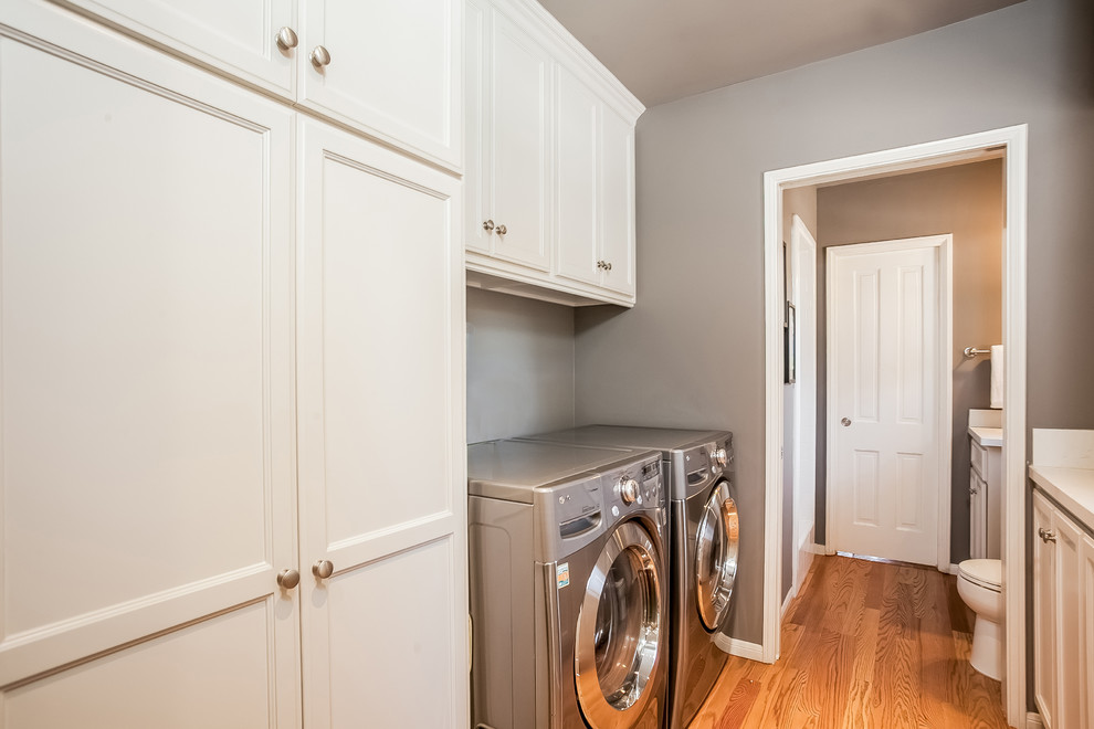 Large transitional l-shaped laundry room in Los Angeles with an undermount sink, shaker cabinets, white cabinets, grey splashback, subway tile splashback, light hardwood floors, quartzite benchtops, grey walls and a side-by-side washer and dryer.