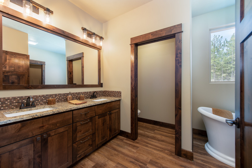 Photo of a mid-sized country master bathroom in Other with beaded inset cabinets, dark wood cabinets, a freestanding tub, brown tile, glass tile, beige walls, medium hardwood floors, an undermount sink, granite benchtops, brown floor and brown benchtops.