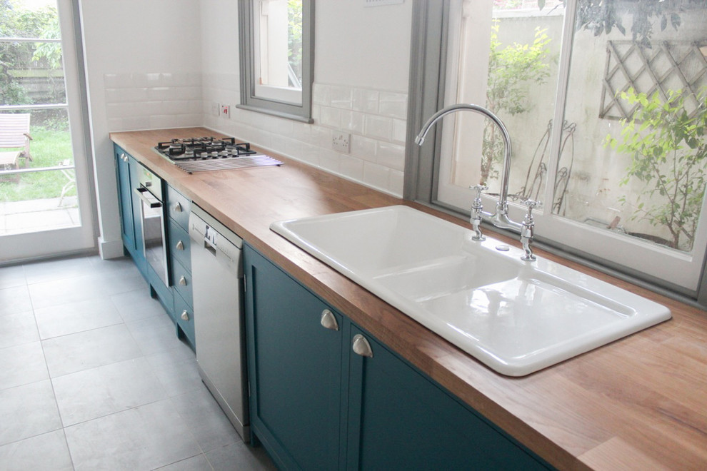 Photo of a small transitional single-wall separate kitchen in London with a double-bowl sink, shaker cabinets, blue cabinets, wood benchtops, white splashback, ceramic splashback, stainless steel appliances and no island.