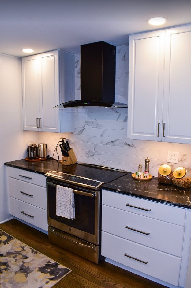 Photo of a mid-sized transitional single-wall eat-in kitchen in Louisville with a farmhouse sink, raised-panel cabinets, white cabinets, quartz benchtops, white splashback, porcelain splashback, black appliances, medium hardwood floors, with island, brown floor and black benchtop.
