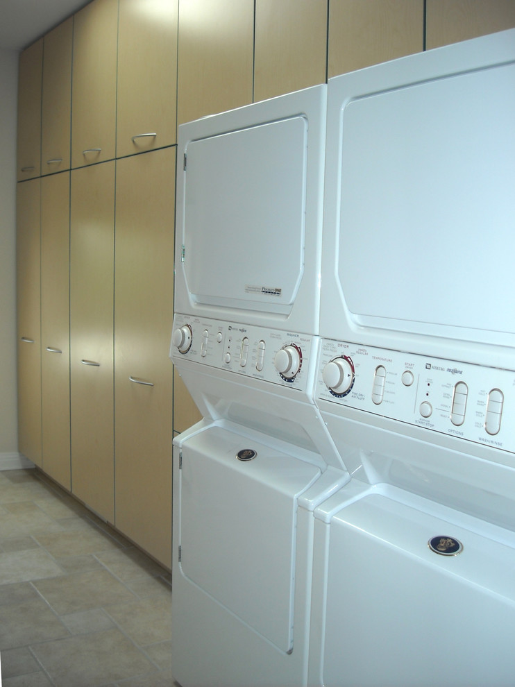 Inspiration for a modern galley laundry room in Chicago with a drop-in sink, flat-panel cabinets, light wood cabinets, laminate benchtops, beige walls, ceramic floors, a stacked washer and dryer and beige floor.