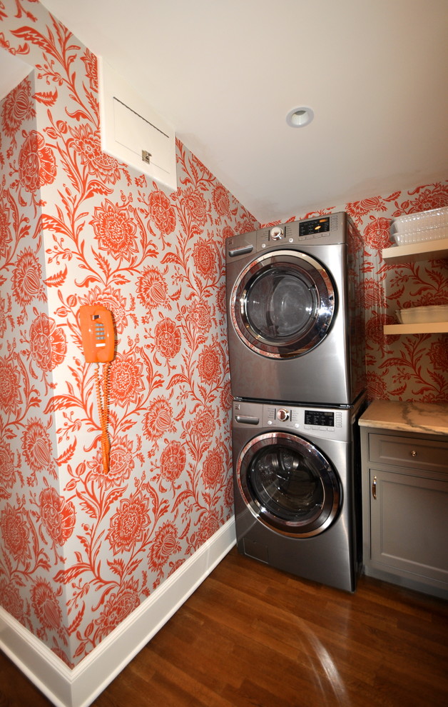 Mid-sized transitional l-shaped utility room in New York with shaker cabinets, grey cabinets, marble benchtops, orange walls, medium hardwood floors and a stacked washer and dryer.
