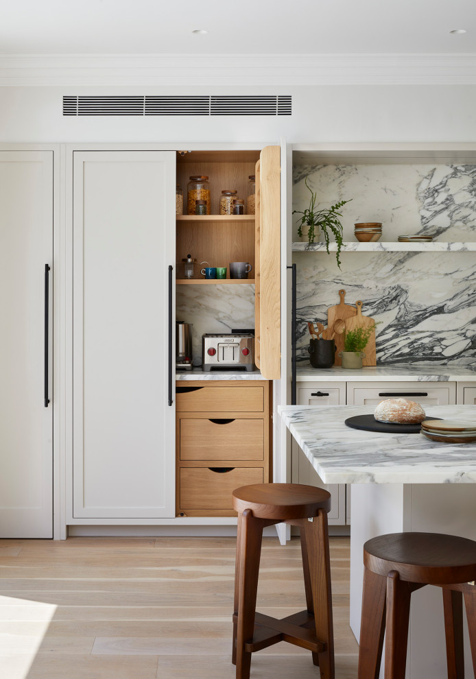 Mid-sized scandinavian l-shaped open plan kitchen in London with a drop-in sink, shaker cabinets, beige cabinets, marble benchtops, white splashback, marble splashback, stainless steel appliances, light hardwood floors, with island and white benchtop.
