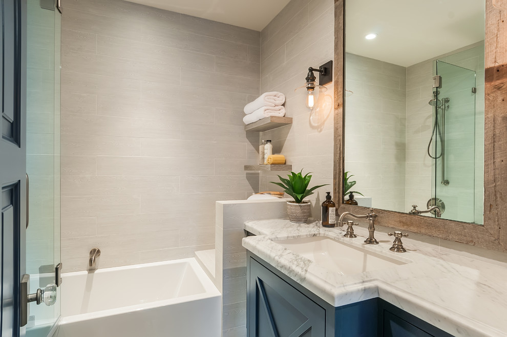 Inspiration for a mid-sized country master bathroom in Los Angeles with recessed-panel cabinets, blue cabinets, an alcove tub, a shower/bathtub combo, gray tile, ceramic tile, grey walls, an undermount sink, marble benchtops, a hinged shower door, white benchtops, limestone floors and grey floor.