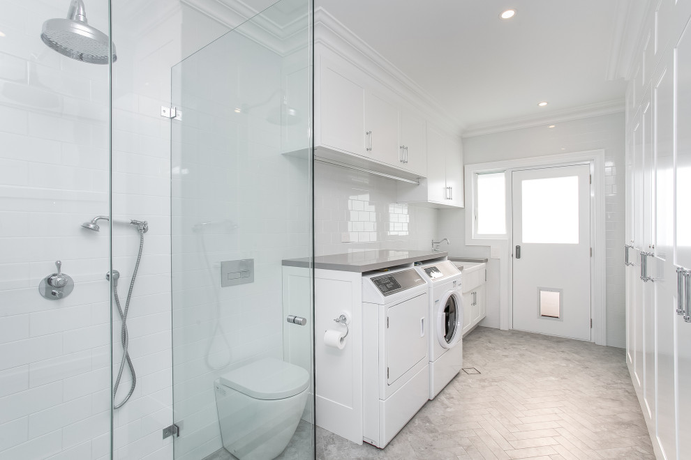Photo of an expansive beach style utility room in Sydney with a farmhouse sink, shaker cabinets, white cabinets, quartz benchtops, white splashback, ceramic splashback, white walls, ceramic floors, a side-by-side washer and dryer, grey floor and grey benchtop.