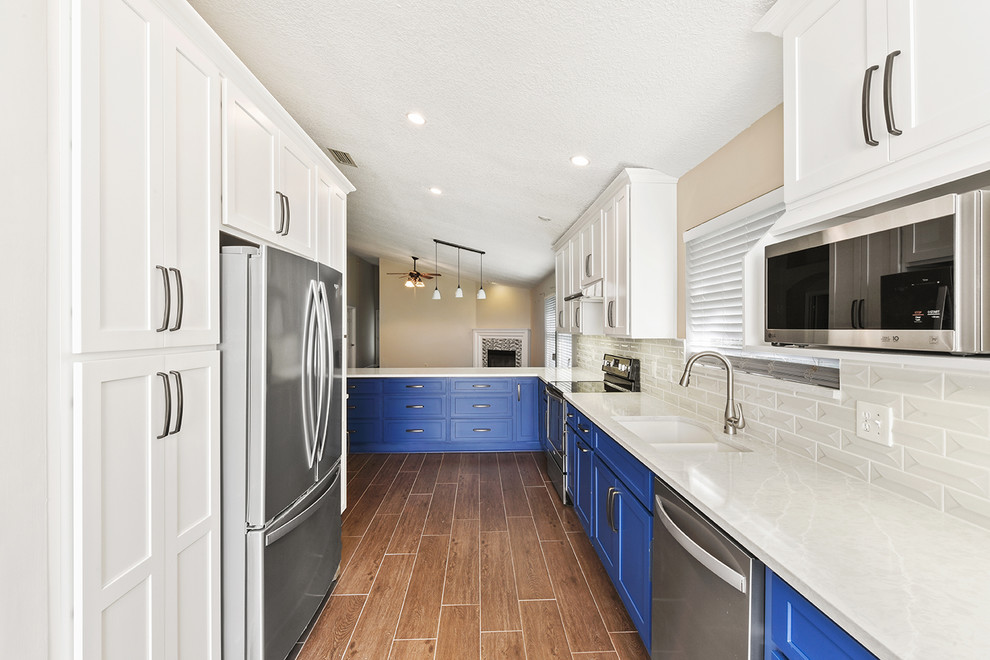 Large transitional l-shaped vinyl floor and brown floor open concept kitchen photo in Jacksonville with an undermount sink, shaker cabinets, blue cabinets, quartzite countertops, white backsplash, ceramic backsplash, stainless steel appliances and a peninsula