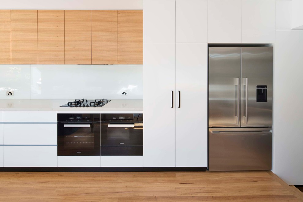 This is an example of a modern single-wall eat-in kitchen in Sydney with an undermount sink, flat-panel cabinets, quartz benchtops, white splashback, glass sheet splashback, black appliances, medium hardwood floors and no island.