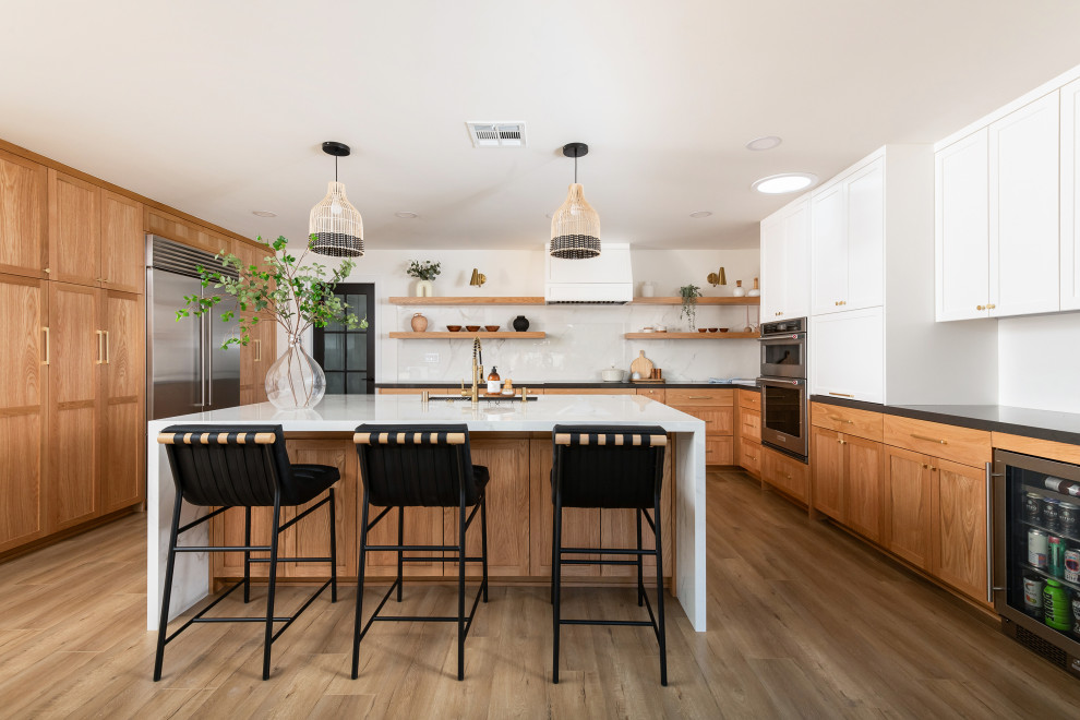 Large beach style l-shaped eat-in kitchen in Orange County with a drop-in sink, shaker cabinets, medium wood cabinets, quartzite benchtops, multi-coloured splashback, stone slab splashback, stainless steel appliances, light hardwood floors, with island and white benchtop.