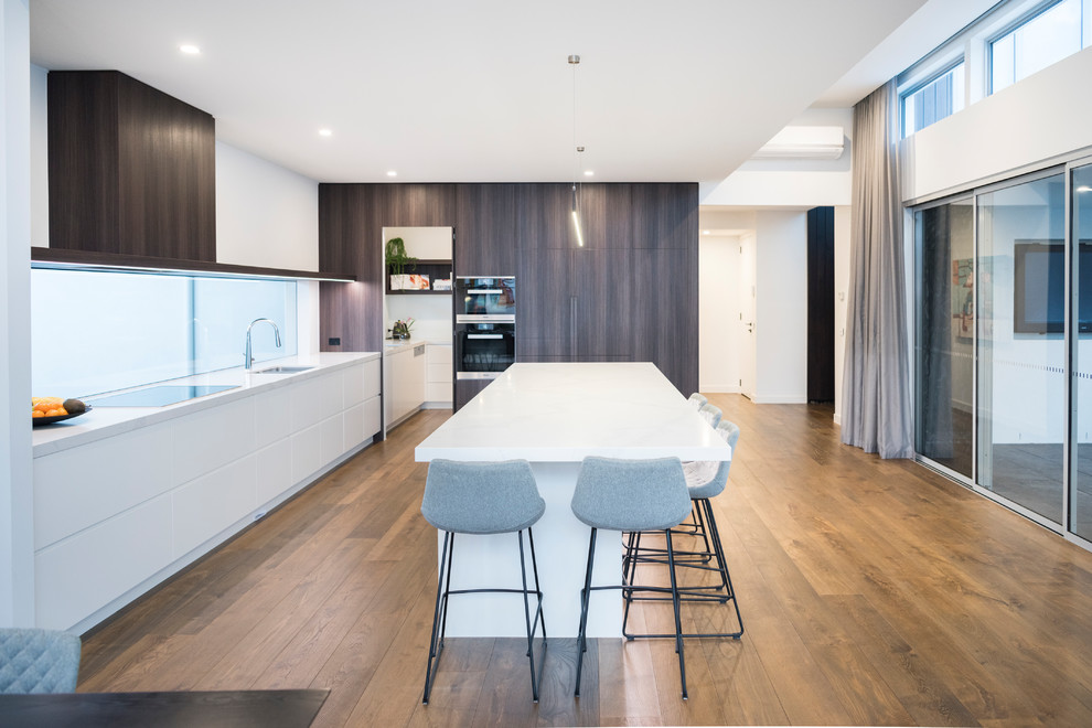 Photo of an expansive contemporary l-shaped open plan kitchen in Melbourne with an undermount sink, quartz benchtops, black appliances, medium hardwood floors, a peninsula, brown floor, white benchtop, flat-panel cabinets, dark wood cabinets and window splashback.