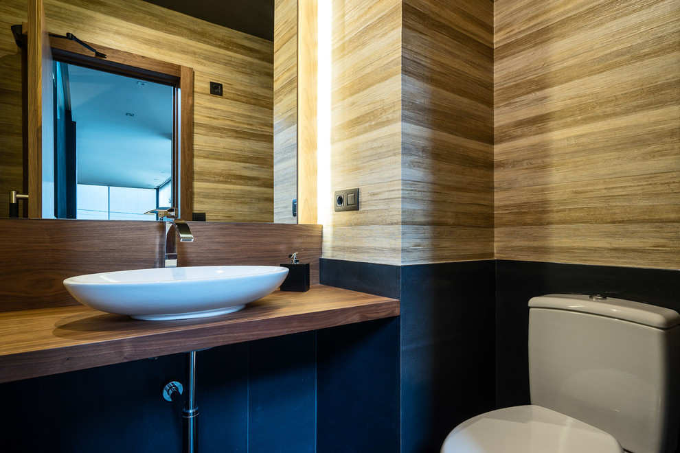 Design ideas for a small modern powder room in Madrid with black tile, porcelain tile, beige walls, porcelain floors, a vessel sink, wood benchtops, black floor, brown benchtops, open cabinets, medium wood cabinets and a two-piece toilet.