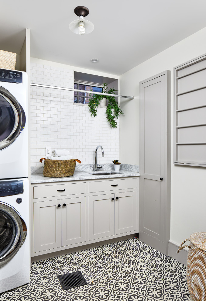 Design ideas for a transitional single-wall dedicated laundry room in Los Angeles with an undermount sink, shaker cabinets, beige cabinets, marble benchtops, white walls, ceramic floors, a stacked washer and dryer, multi-coloured floor and grey benchtop.