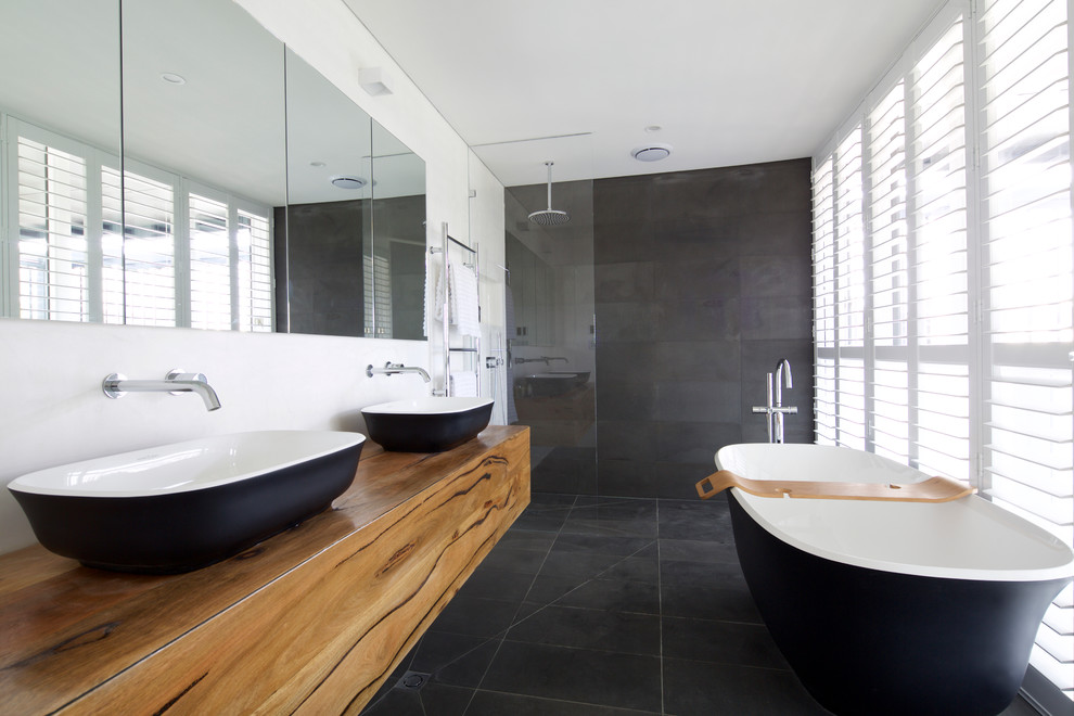 This is an example of an expansive contemporary master bathroom in Sydney with a vessel sink, flat-panel cabinets, medium wood cabinets, wood benchtops, a freestanding tub, an open shower, gray tile, stone tile, white walls, slate floors, black floor, an open shower and brown benchtops.