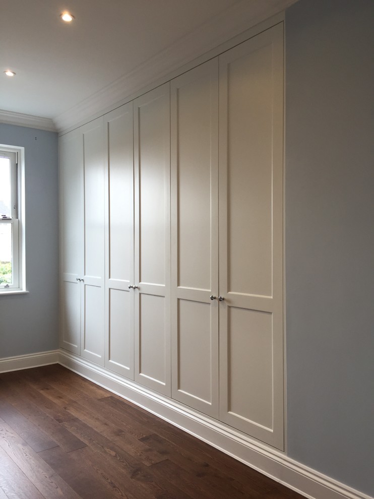 This is an example of a large traditional gender-neutral built-in wardrobe in London with recessed-panel cabinets, white cabinets, dark hardwood floors and brown floor.