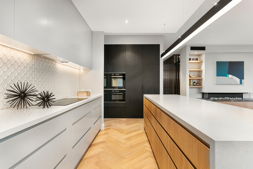 This is an example of a contemporary u-shaped open plan kitchen in Melbourne with flat-panel cabinets, light wood cabinets, concrete benchtops, white splashback, black appliances, light hardwood floors, with island, beige floor and grey benchtop.