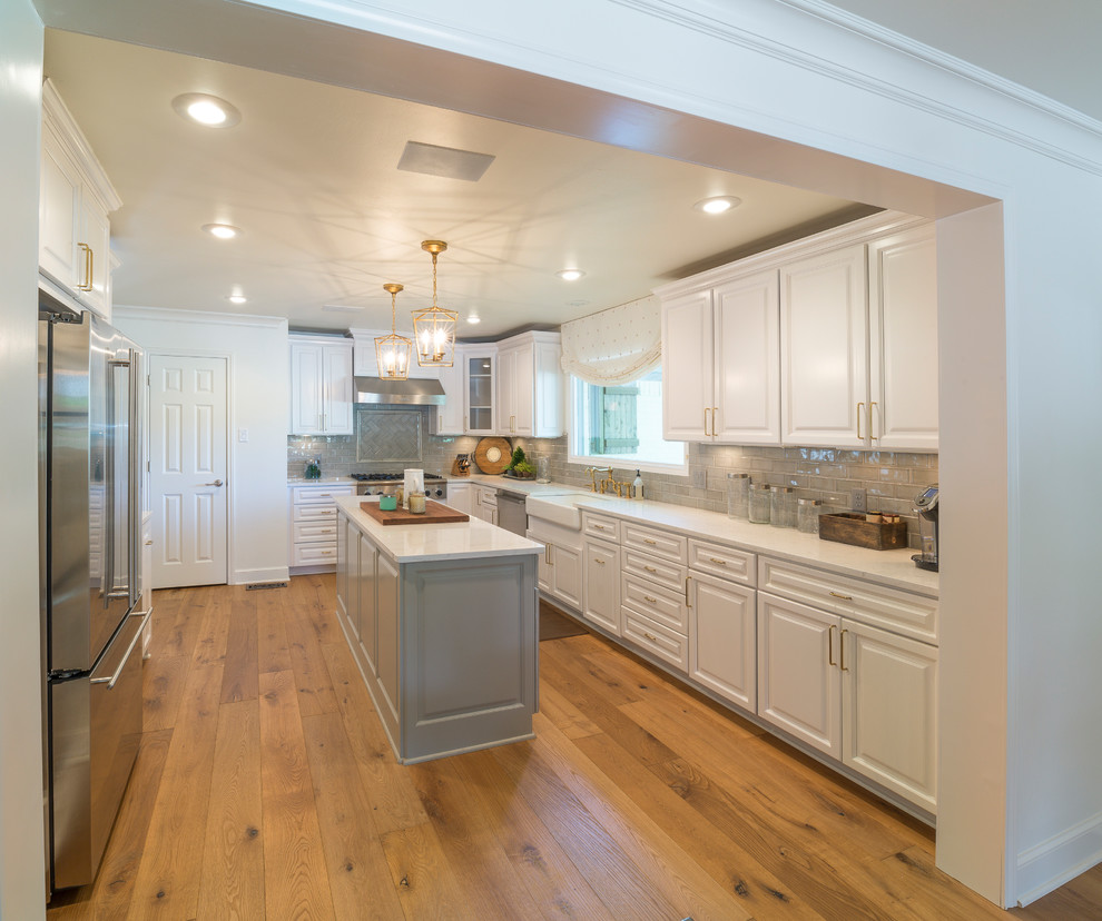 Large transitional u-shaped medium tone wood floor and brown floor enclosed kitchen photo with a farmhouse sink, raised-panel cabinets, white cabinets, quartz countertops, gray backsplash, glass tile backsplash, stainless steel appliances and an island