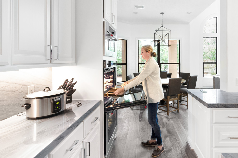 Inspiration for a mid-sized transitional galley medium tone wood floor and gray floor eat-in kitchen remodel in Houston with a farmhouse sink, raised-panel cabinets, blue cabinets, quartzite countertops, gray backsplash, stone slab backsplash, stainless steel appliances, an island and gray countertops