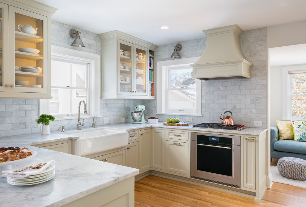 This is an example of a mid-sized transitional u-shaped eat-in kitchen in Minneapolis with a farmhouse sink, flat-panel cabinets, marble benchtops, grey splashback, marble splashback, stainless steel appliances, light hardwood floors, a peninsula, brown floor, grey benchtop and beige cabinets.