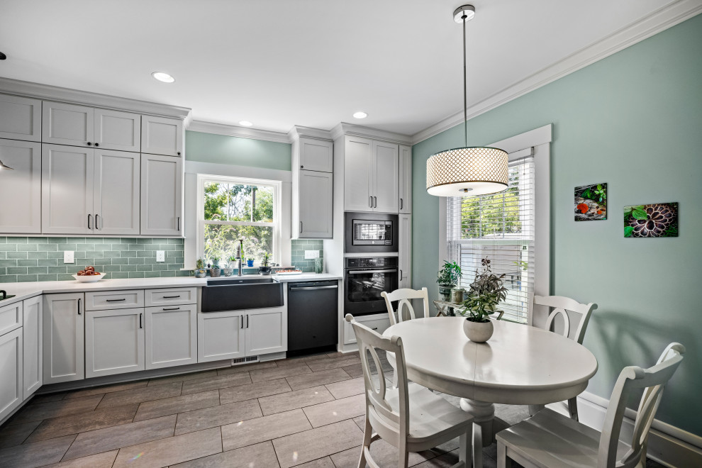 Mid-sized traditional l-shaped eat-in kitchen in Columbus with a farmhouse sink, shaker cabinets, white cabinets, quartz benchtops, green splashback, cement tile splashback, black appliances, porcelain floors, a peninsula, grey floor and white benchtop.