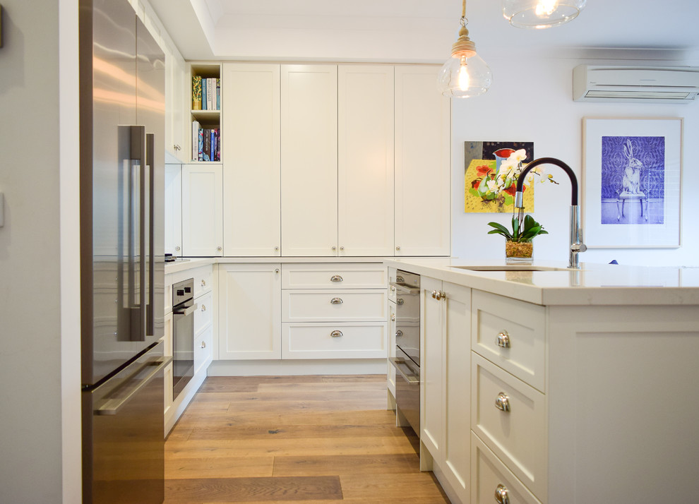 Design ideas for a mid-sized transitional l-shaped open plan kitchen in Melbourne with an undermount sink, shaker cabinets, white cabinets, quartz benchtops, white splashback, subway tile splashback, stainless steel appliances, light hardwood floors, with island and brown floor.