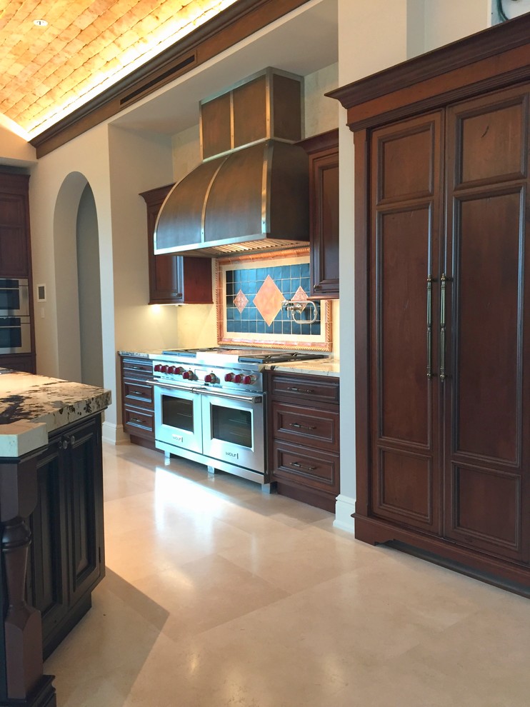 This is an example of an expansive mediterranean open plan kitchen in Miami with a farmhouse sink, beaded inset cabinets, dark wood cabinets, granite benchtops, multi-coloured splashback, ceramic splashback, panelled appliances, travertine floors, with island, beige floor and multi-coloured benchtop.